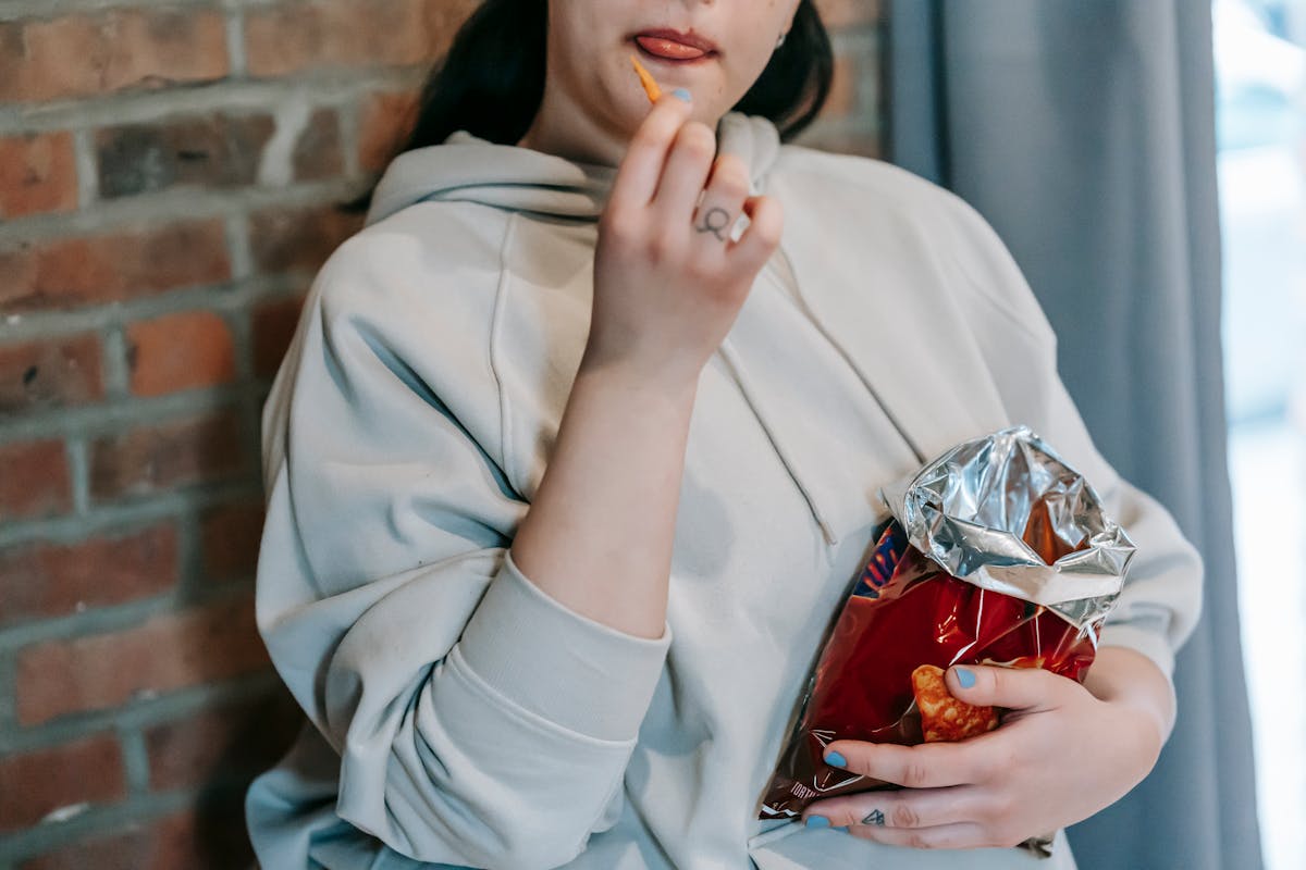 Crop plump female in casual hoodie enjoying crispy baked breads and licking lips against brick wall snack