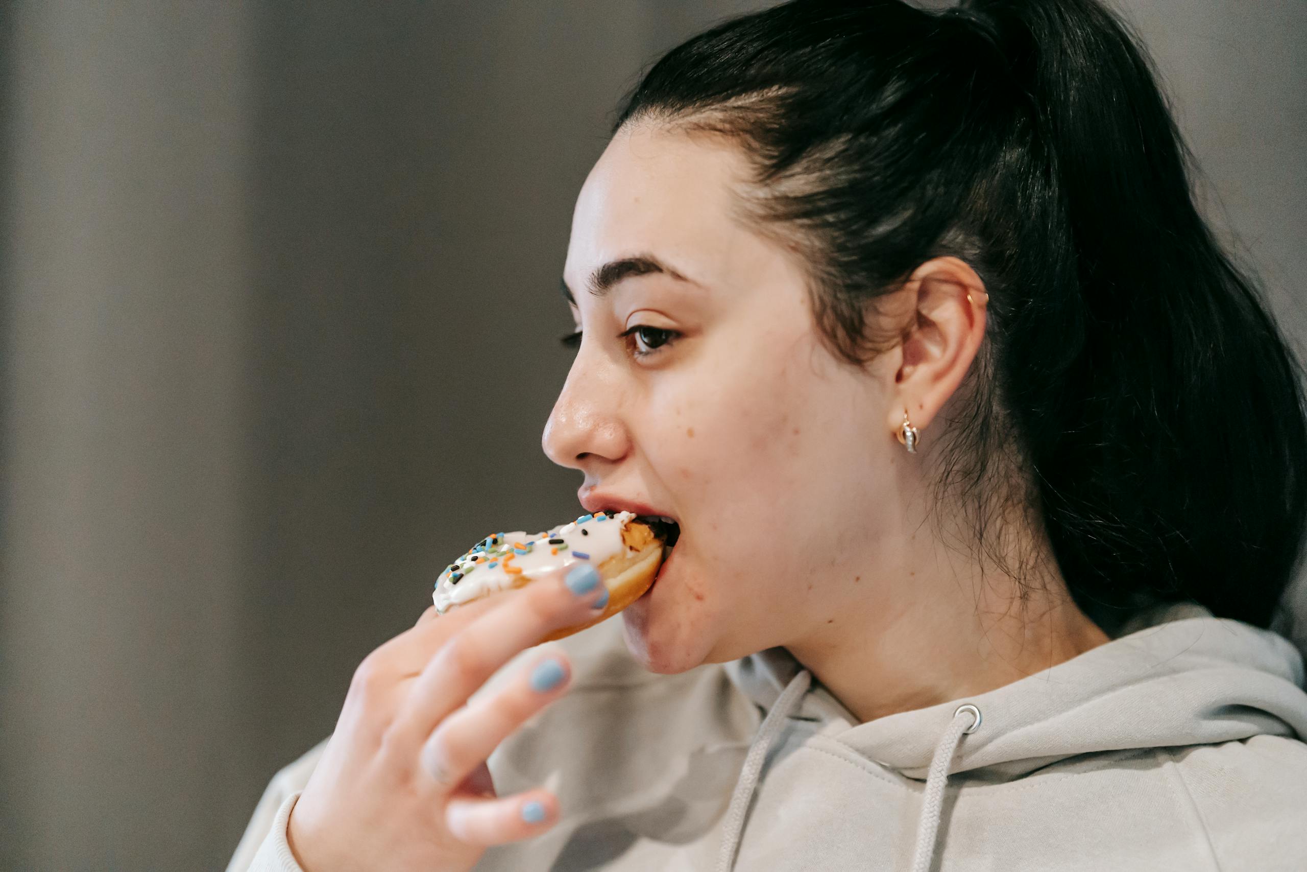 Crop young brunette in comfy hoodie eating fresh appetizing doughnut covered with vanilla icing and sprinkles in light room
