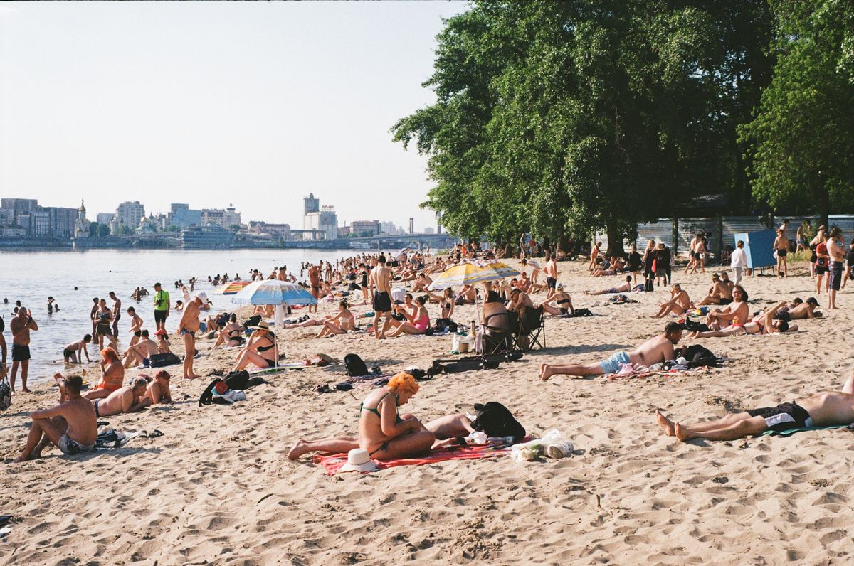 Crowded beach scene with people enjoying the sand and sun under green trees.