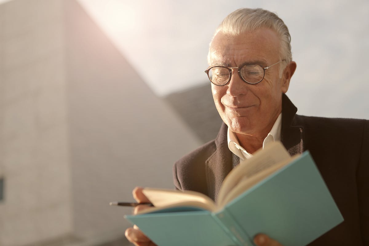 12 Routines Retirees Can Incorporate Into Their Lives To Help Kill Boredom 2 Elderly man wearing glasses, reading a book outdoors with a smile on a sunny day.