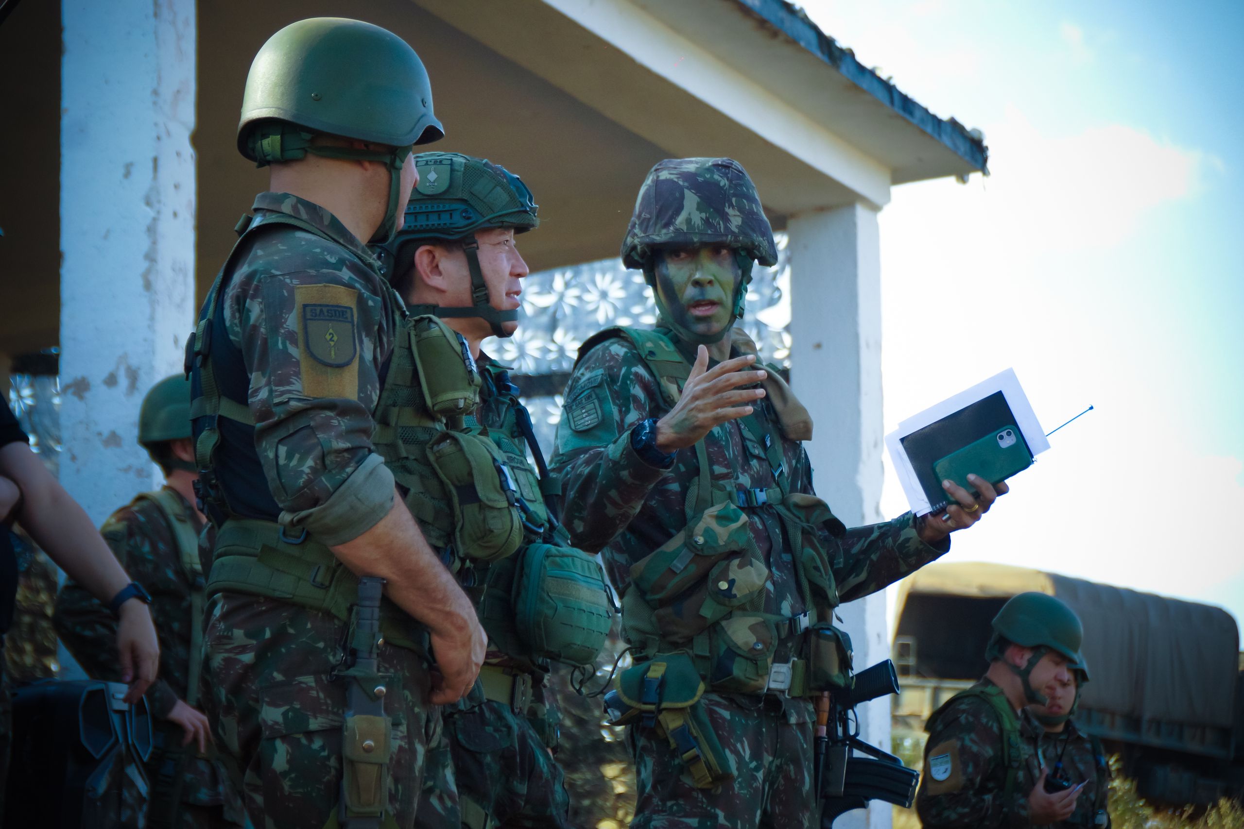 Group of soldiers in camouflage attending a briefing outdoors with tactical gear and helmets.