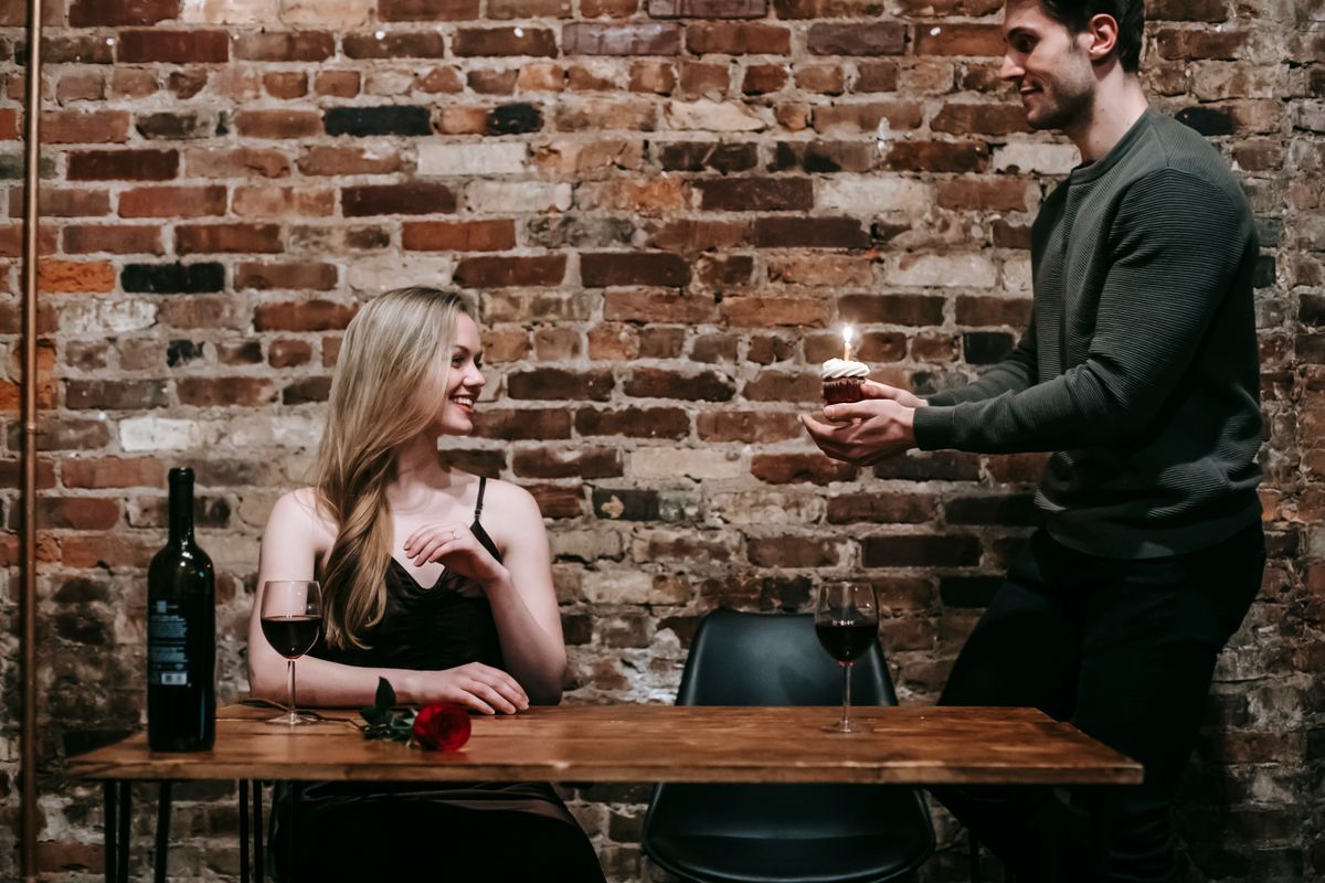 Happy young man giving woman birthday cupcake and candle on date in restaurant at table with glasses and bottle with red wine and rose near brick wall