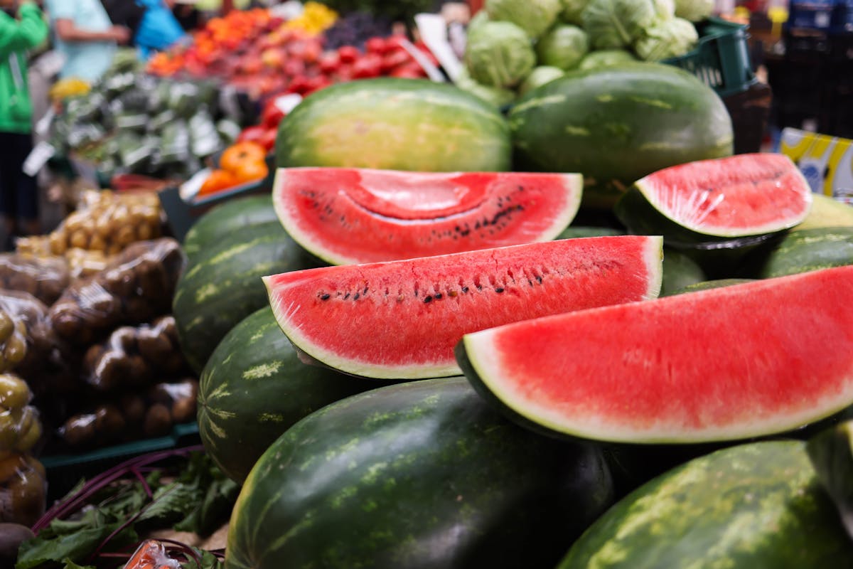 Beware Before You Grab That Watermelon: Shocking Truths Grocery Stores Won’t Tell You 2 Juicy watermelon slices stacked among fresh fruits and vegetables at an outdoor market.