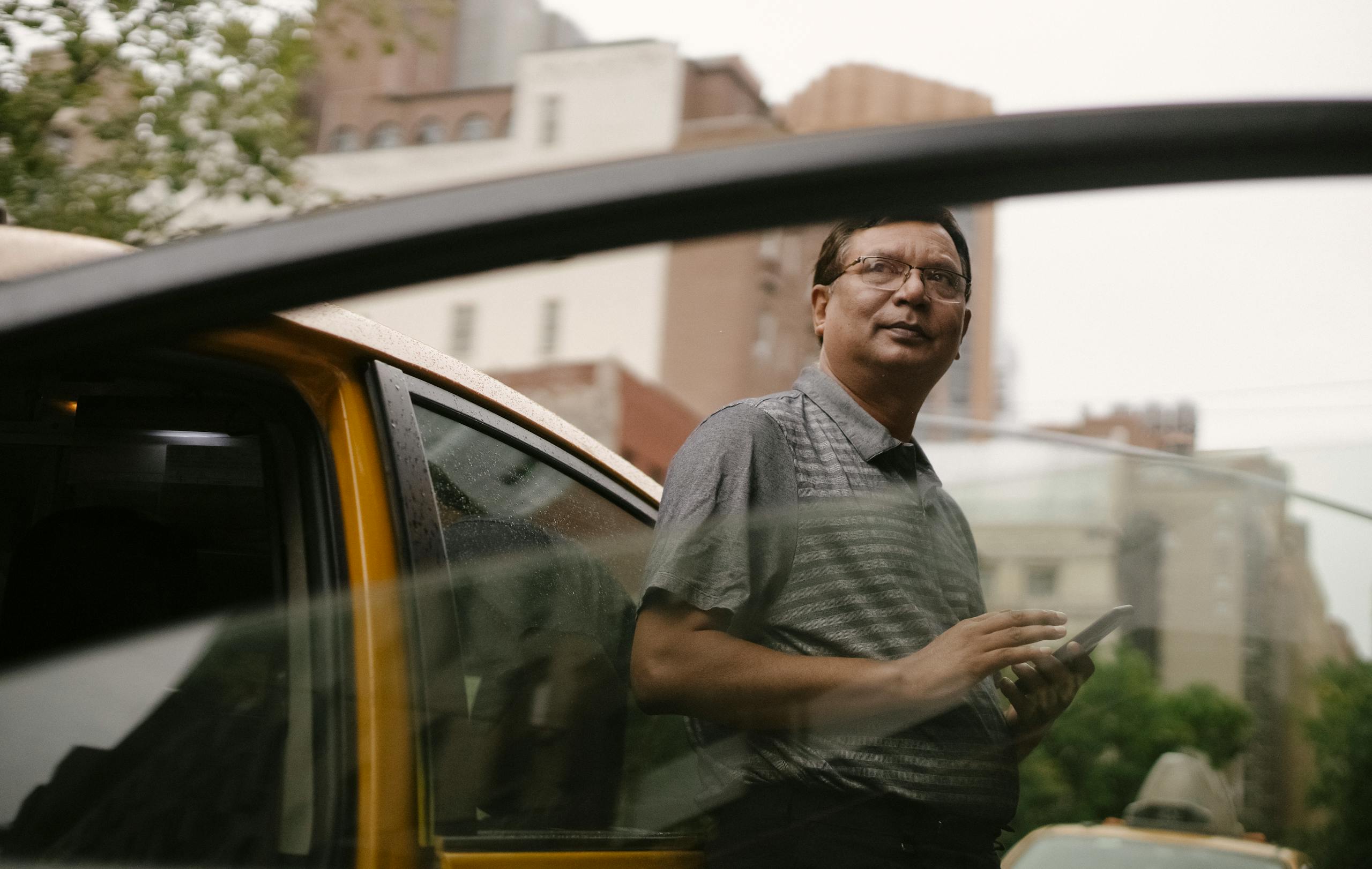 Low angle of pensive male driver in casual clothes leaning on door of cab while holding mobile phone and looking away