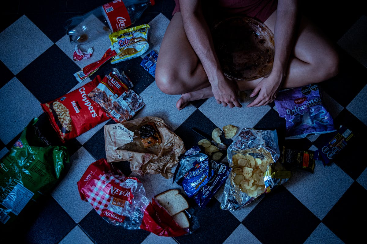 Man sitting surrounded by assorted snacks and packaging on a checkered floor, depicting a binge-eating scene. Late-Night Snacking