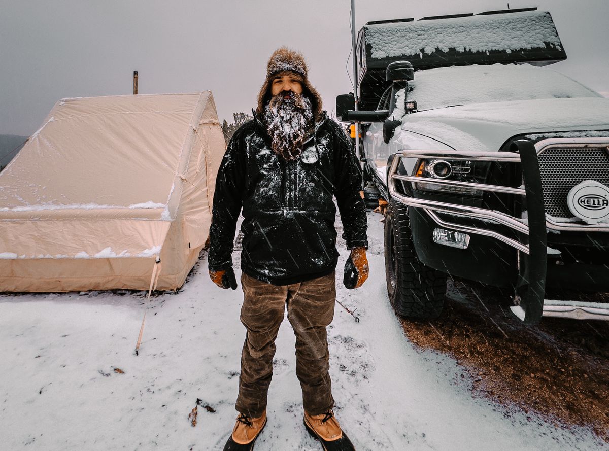 10 Harshest Places People Call Home 2 Man with beard standing next to a tent and truck in a snowy winter scene, suggesting adventure.