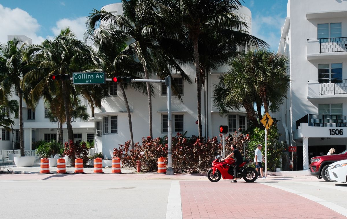 Miami Sellers Are Pulling Homes Off the Market Instead of Cutting Prices, and the Numbers Reveal a Bigger Housing Standoff 2 Motorcycle passing Collins Avenue in sunny Miami Beach, with palm trees and art deco architecture.