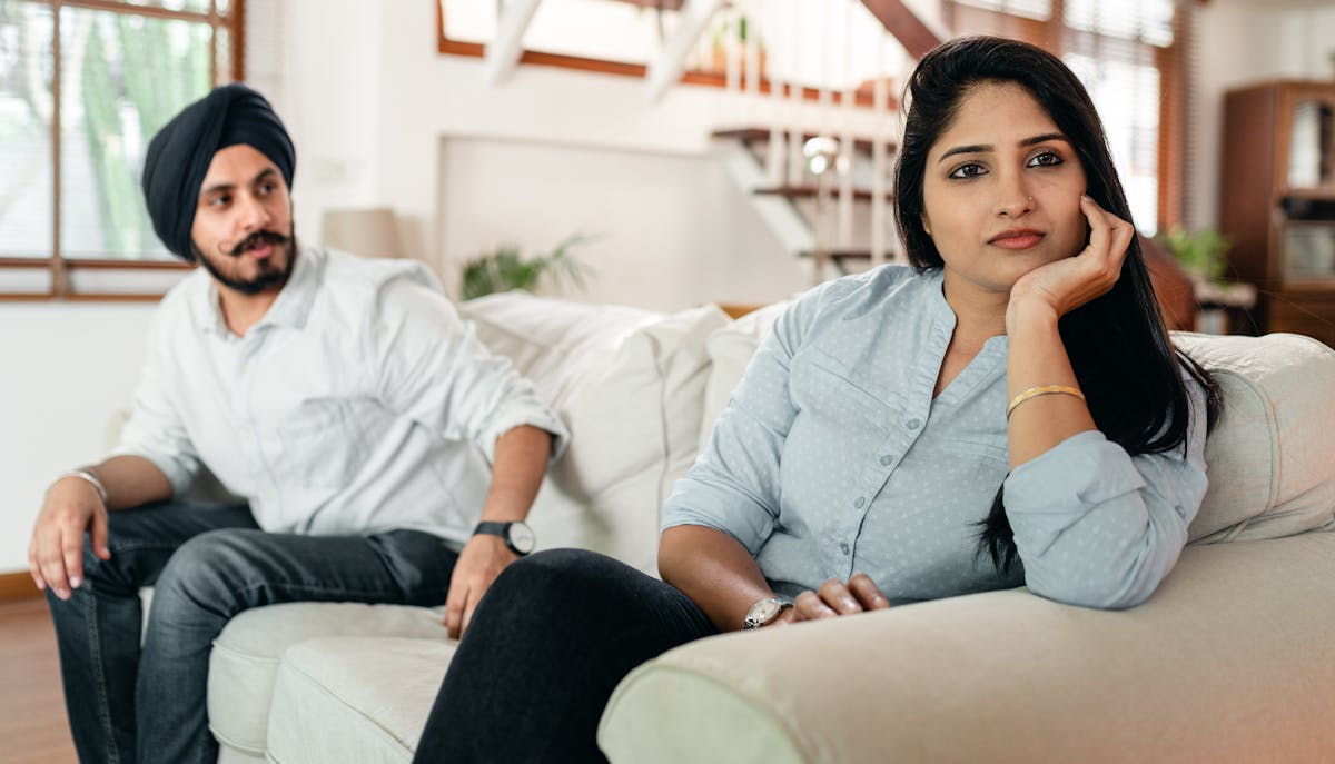 Offended Indian woman sitting on sofa and looking sadly away while bearded husband trying to make peace after quarrel