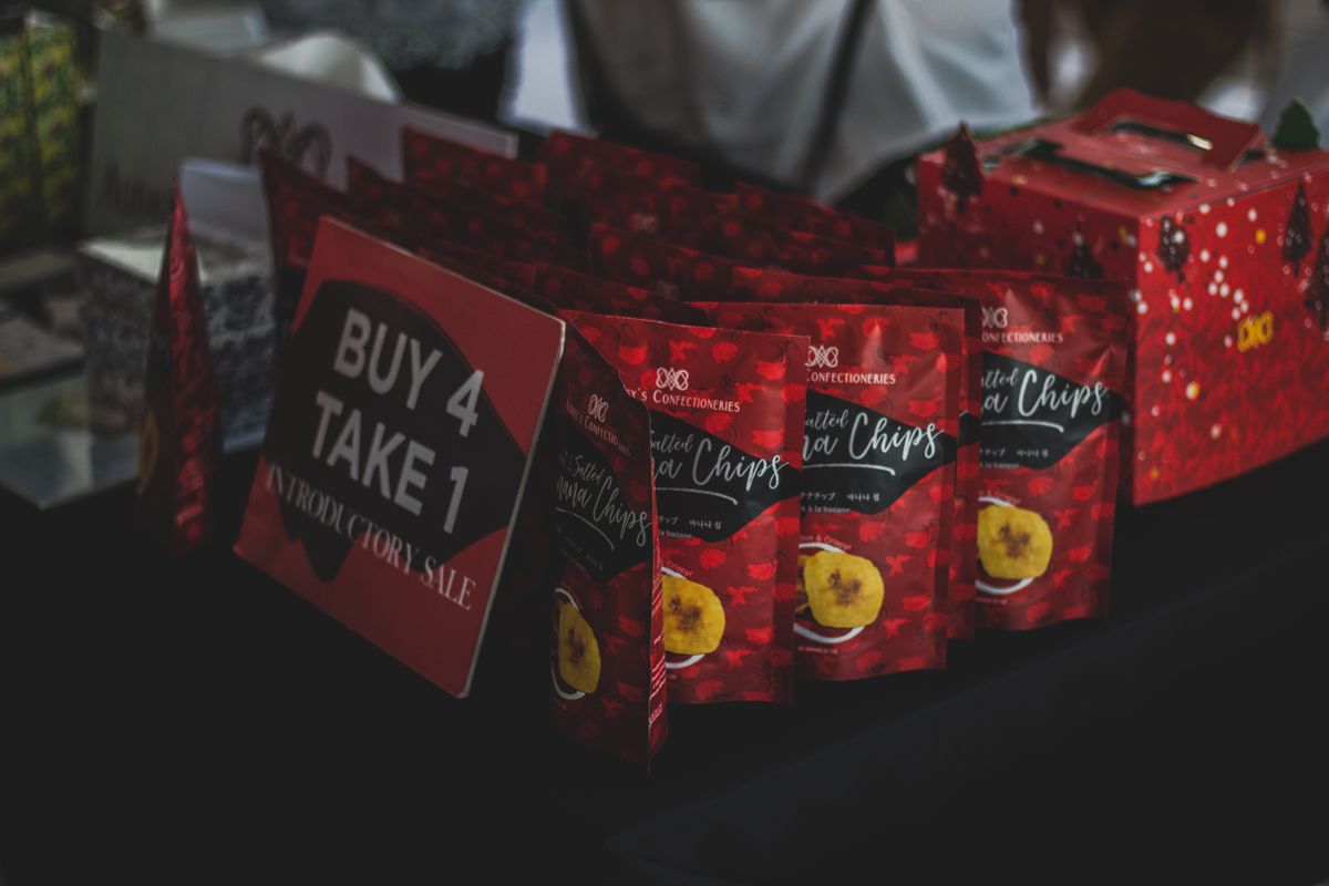 Packaged banana chips displayed with a promotional 'Buy 4 Take 1' sign at an indoor sale event.