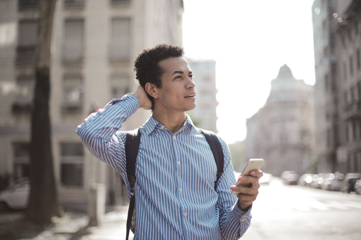 Pensive young ethnic man with smartphone standing on street looking for location being lost while walking in city