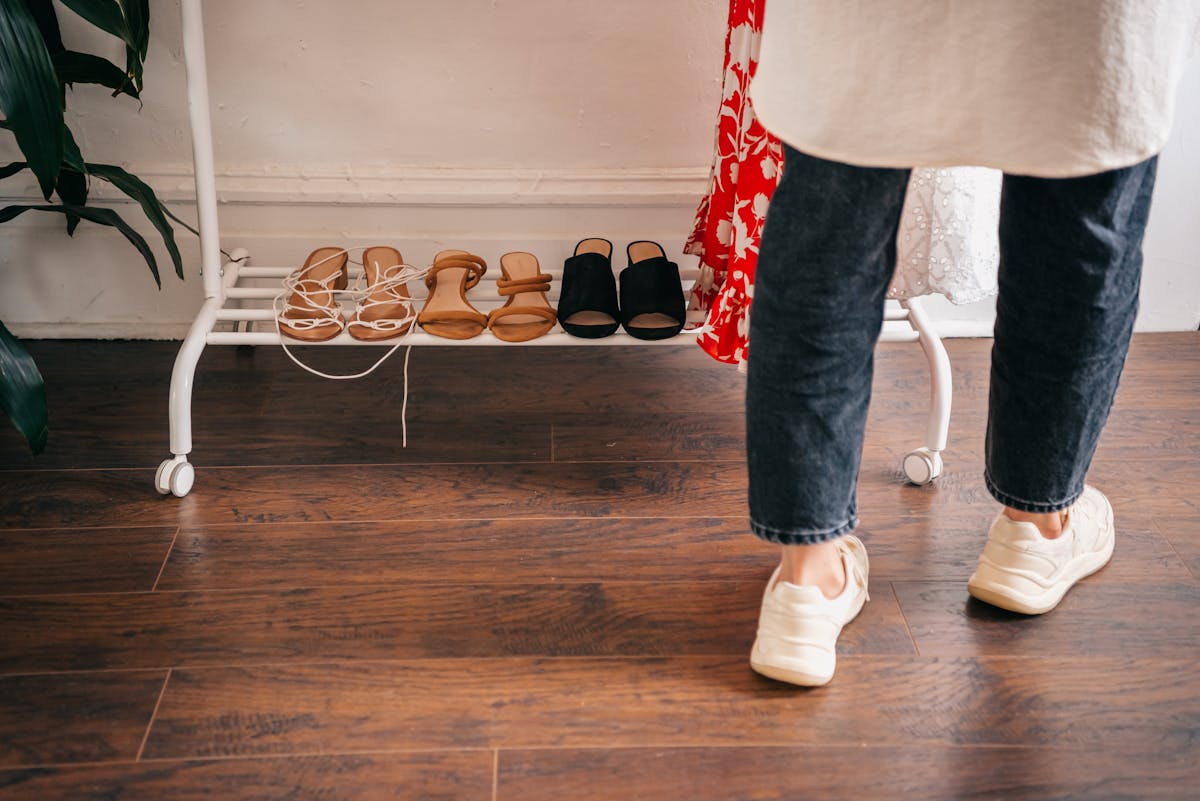 Person in casual attire browsing shoes on a rack indoors, wooden flooring visible.
