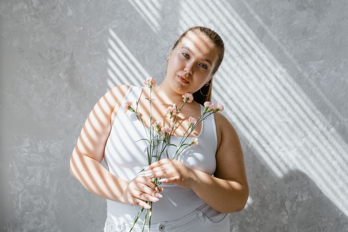 8 Backhanded Compliments Plus-Size Women Are Exhausted From Hearing 2 Plus-size woman in tank top holding flowers with sunlight filtering through blinds.