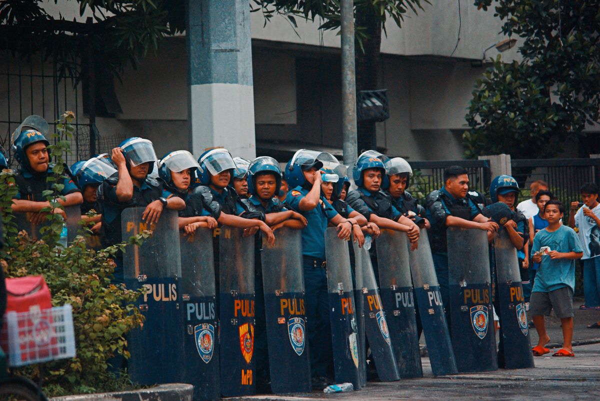 Police officers in riot gear line the street during a protest. Crowd of people observing nearby.