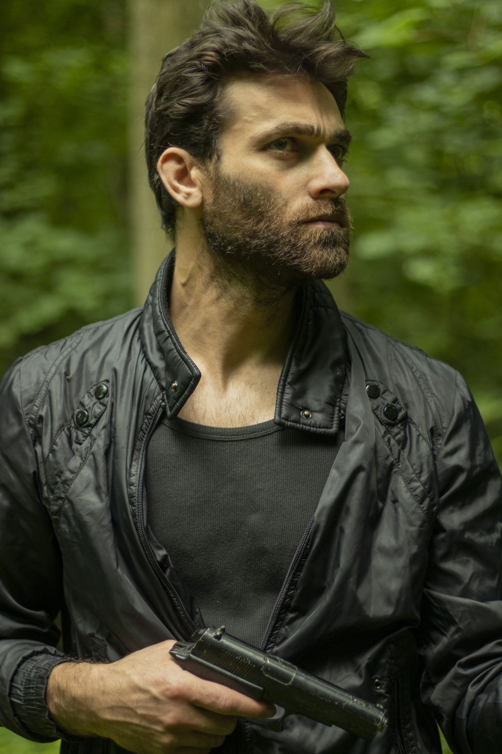 Portrait of a bearded man in a black jacket holding a pistol outdoors.