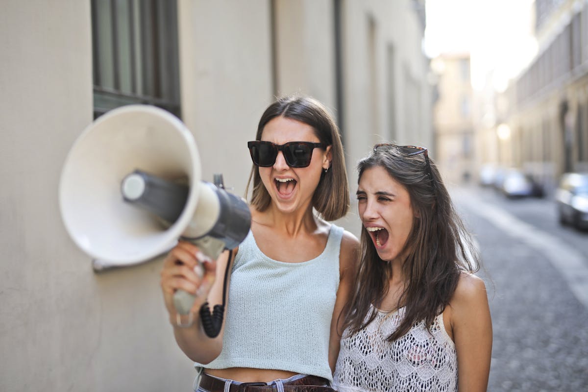 Positive female friends having fun while shouting into megaphone and smiling when standing on city street
