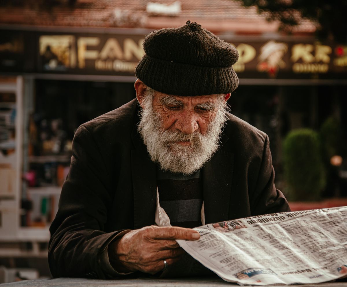 10 hard truths that made boomers mentally tough 1 Senior man in woolen cap reading a newspaper on city streets, captured in a candid street photography style.