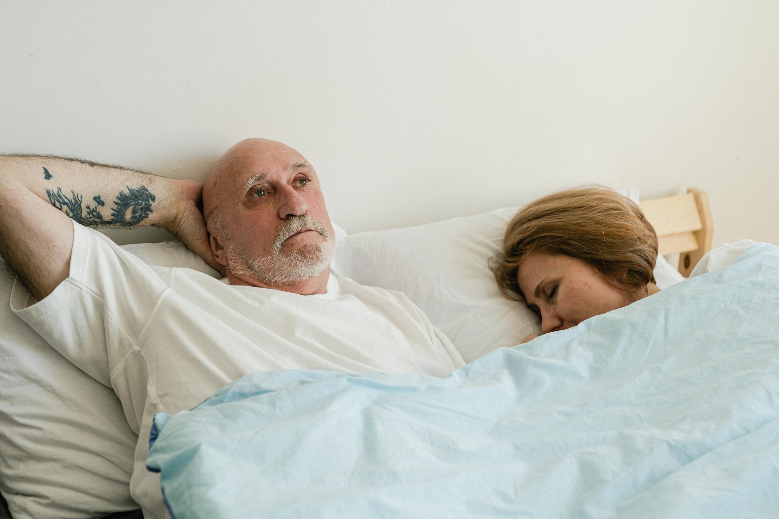 Senior man with tattoo and woman resting in bed, serene indoor setting.