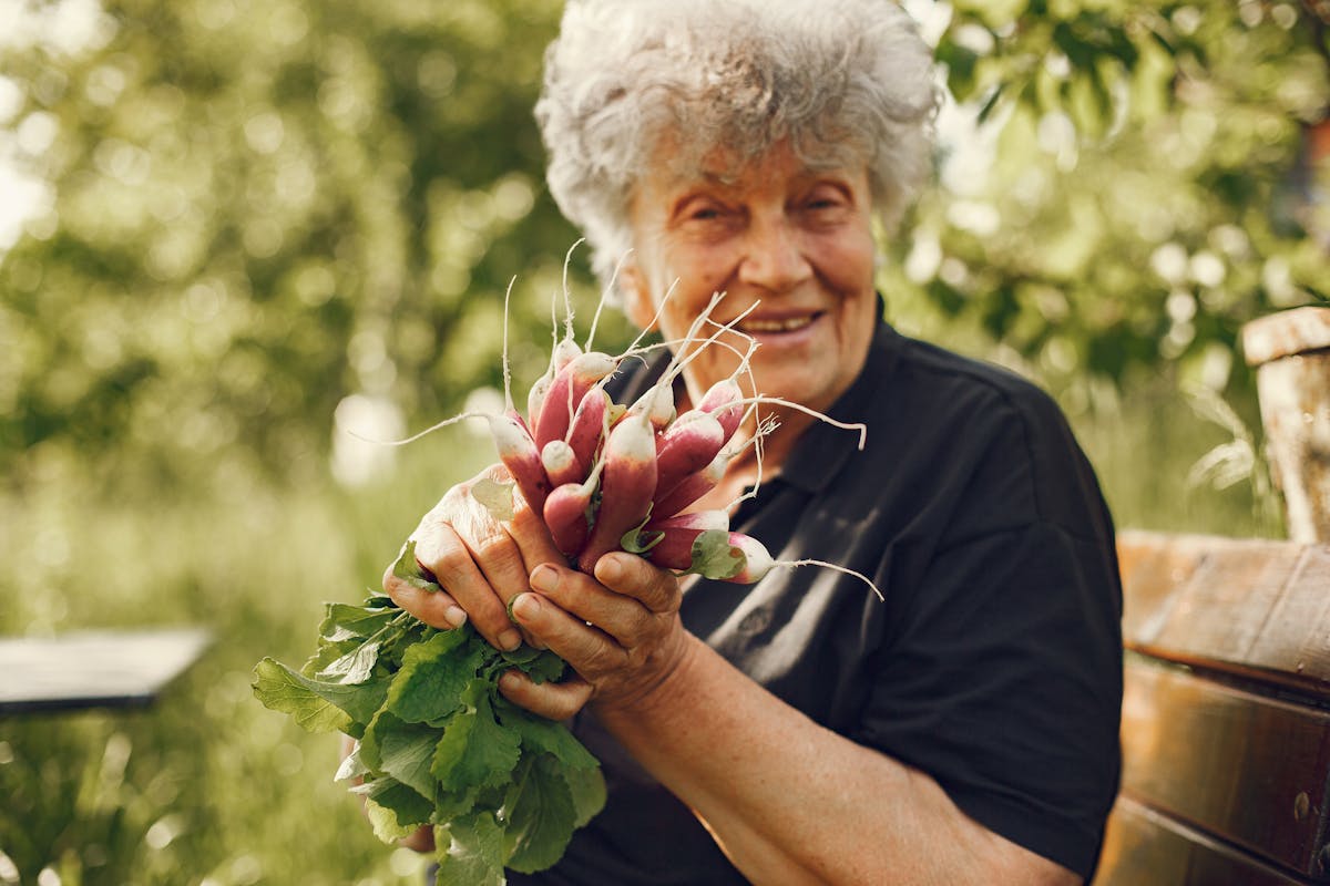Senior woman smiling while holding a bunch of freshly harvested radishes outdoors in a garden.