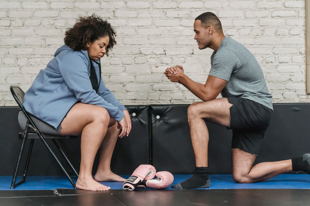 Side view of muscular African American male trainer explaining technique of exercise to plump black woman while sitting on mat with boxing gloves