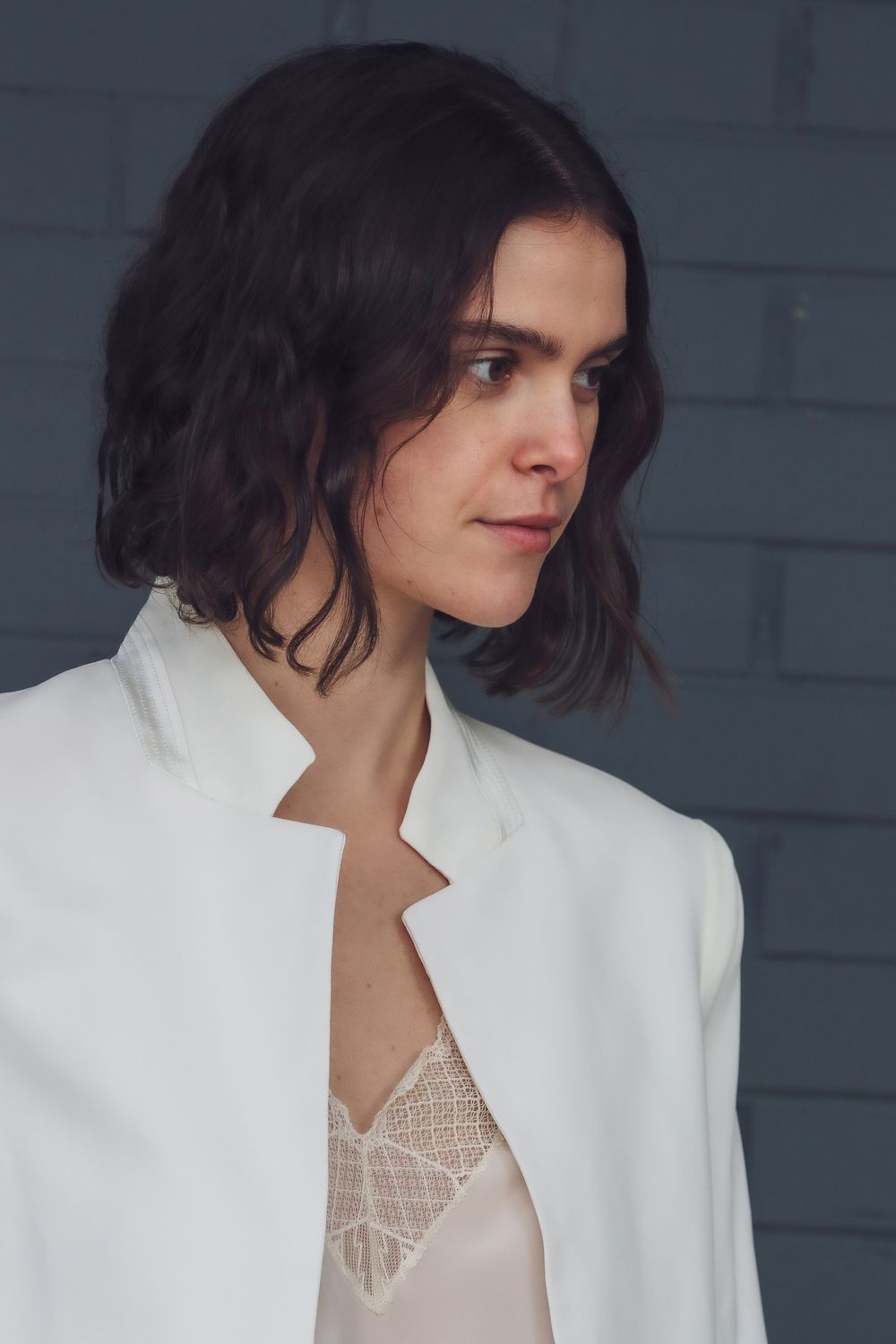 Stylish brunette woman wearing a white blazer against a brick wall.