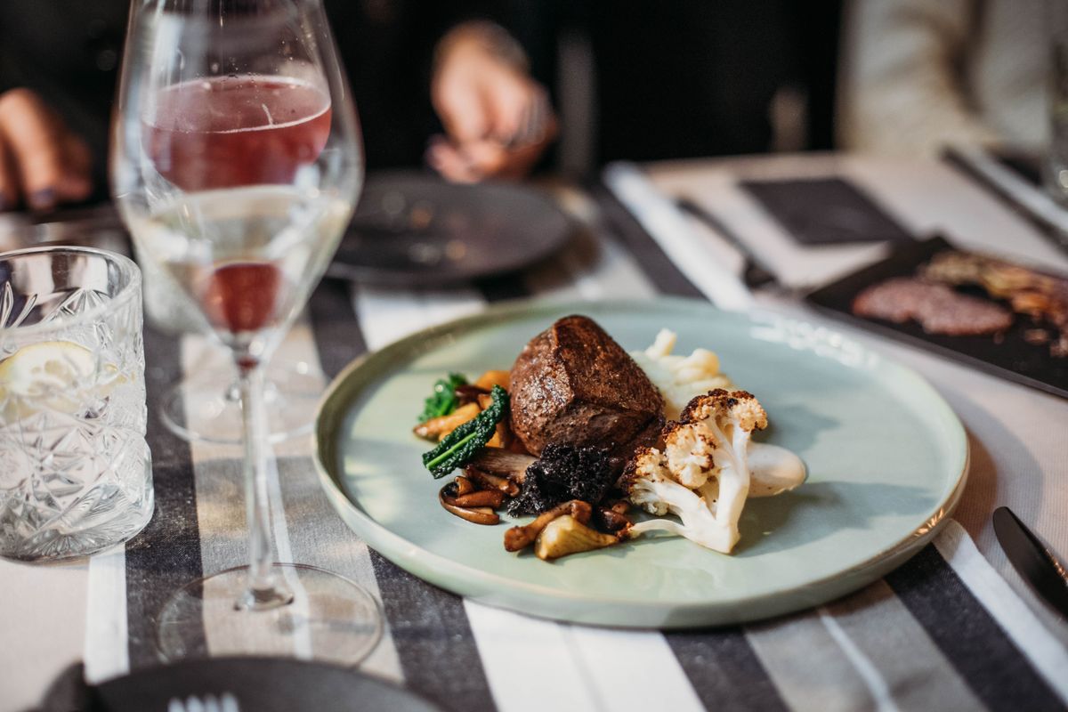 New Katy Restaurant Brings Global Comfort Food, Cocktails, and Wine Tastings To Mason Road 1 Tasty grilled steak served with cauliflower and mushrooms placed on table with wineglass filled with alcohol drink in restaurant on blurred background