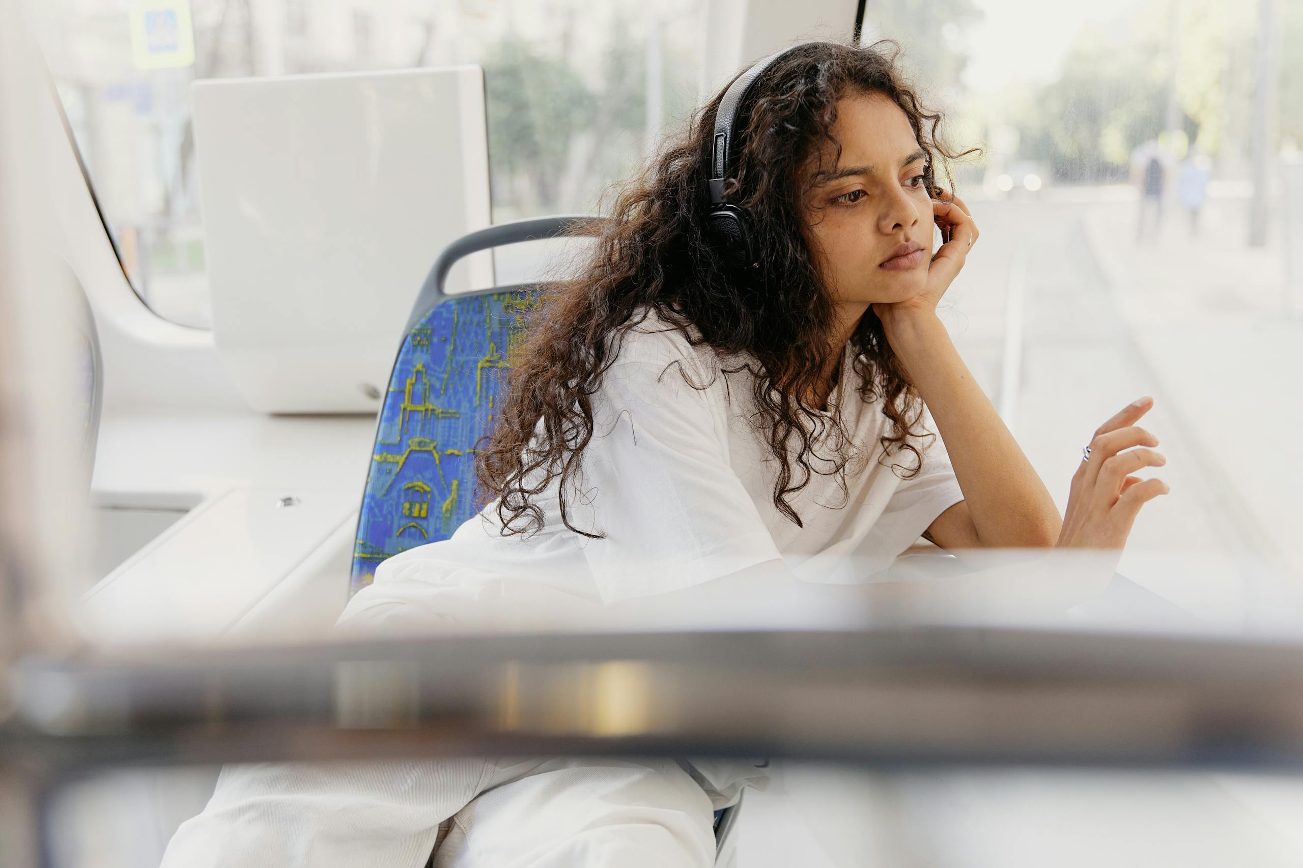 Thoughtful woman with curly hair in white shirt listening to music on train.