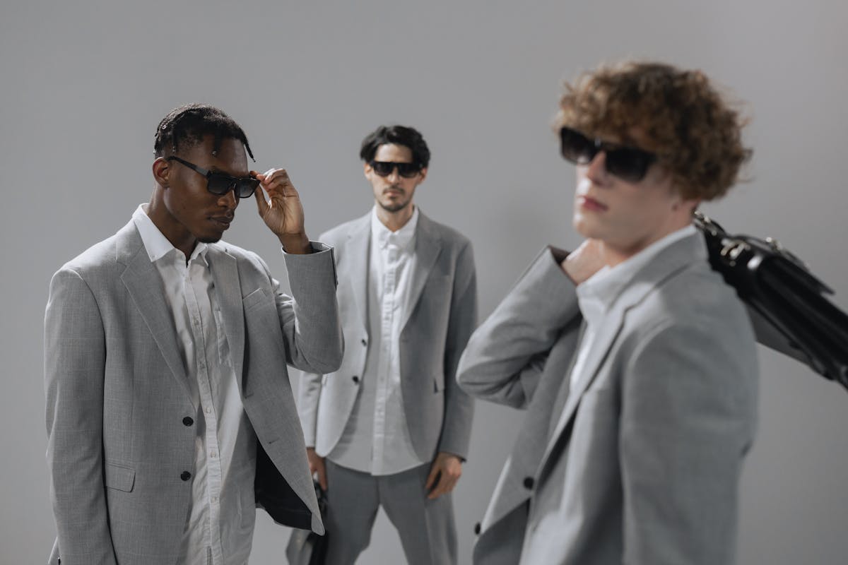 8 Men’s Fashion Choices That Scream “Notice Me Now.” 1 Three stylish men in gray suits and sunglasses exuding professionalism and modern corporate style.