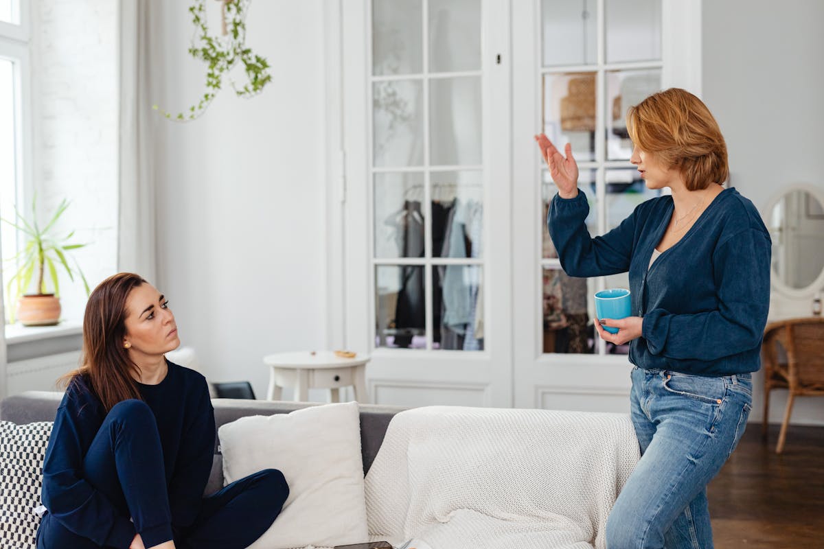 Two women engage in a friendly chat in a stylish living room, one sitting and one standing with a mug.