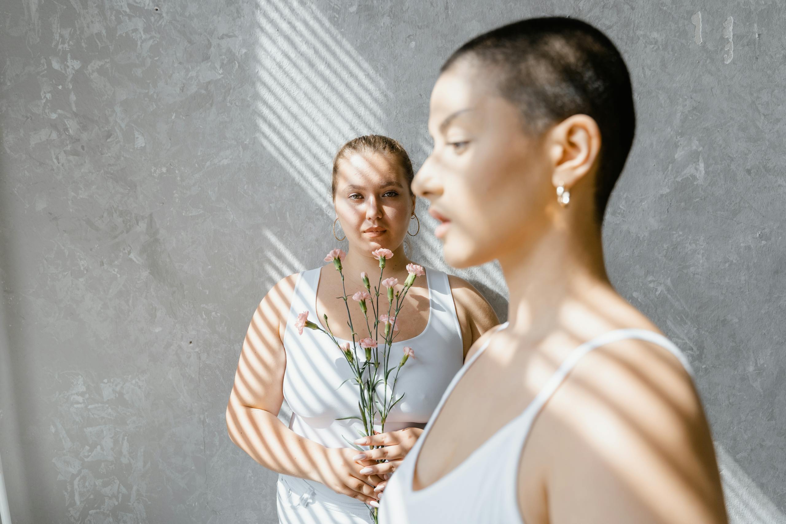 Two women in white tank tops holding flowers in an artistic indoor setting.