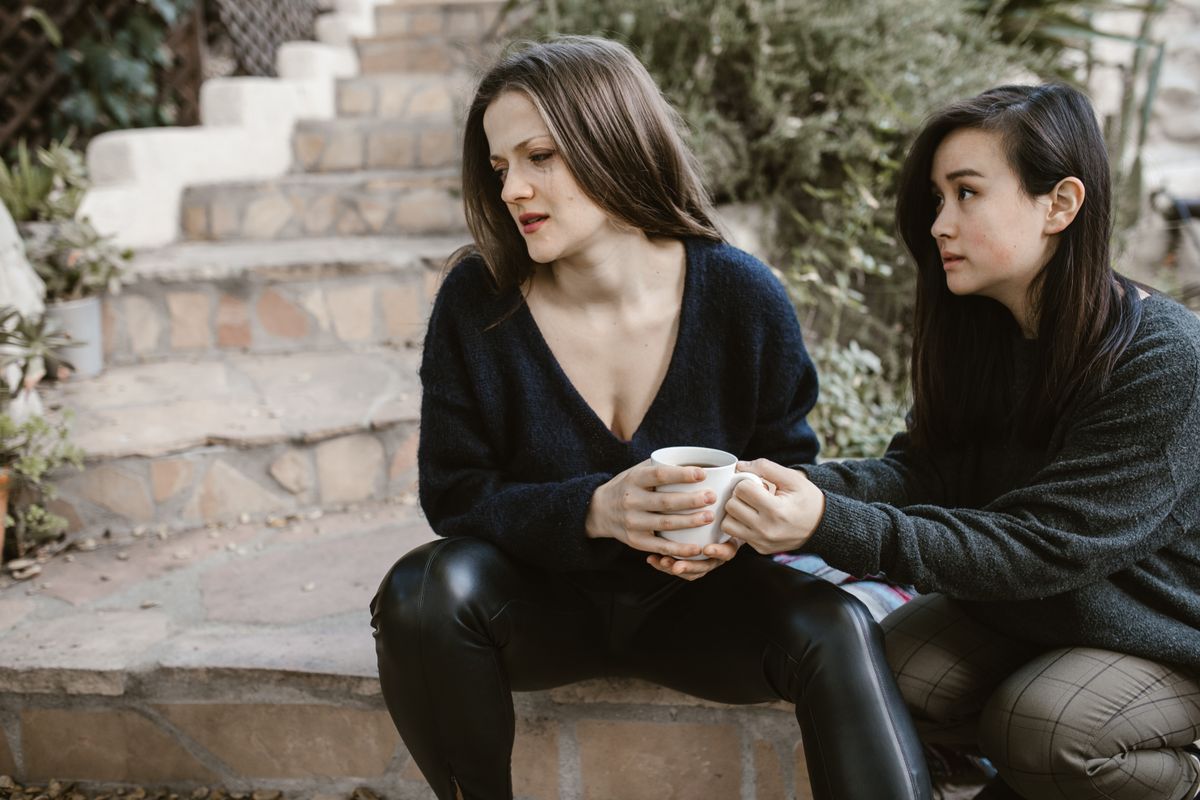 Two women share emotional support on outdoor steps, one consoling the other with a cup.