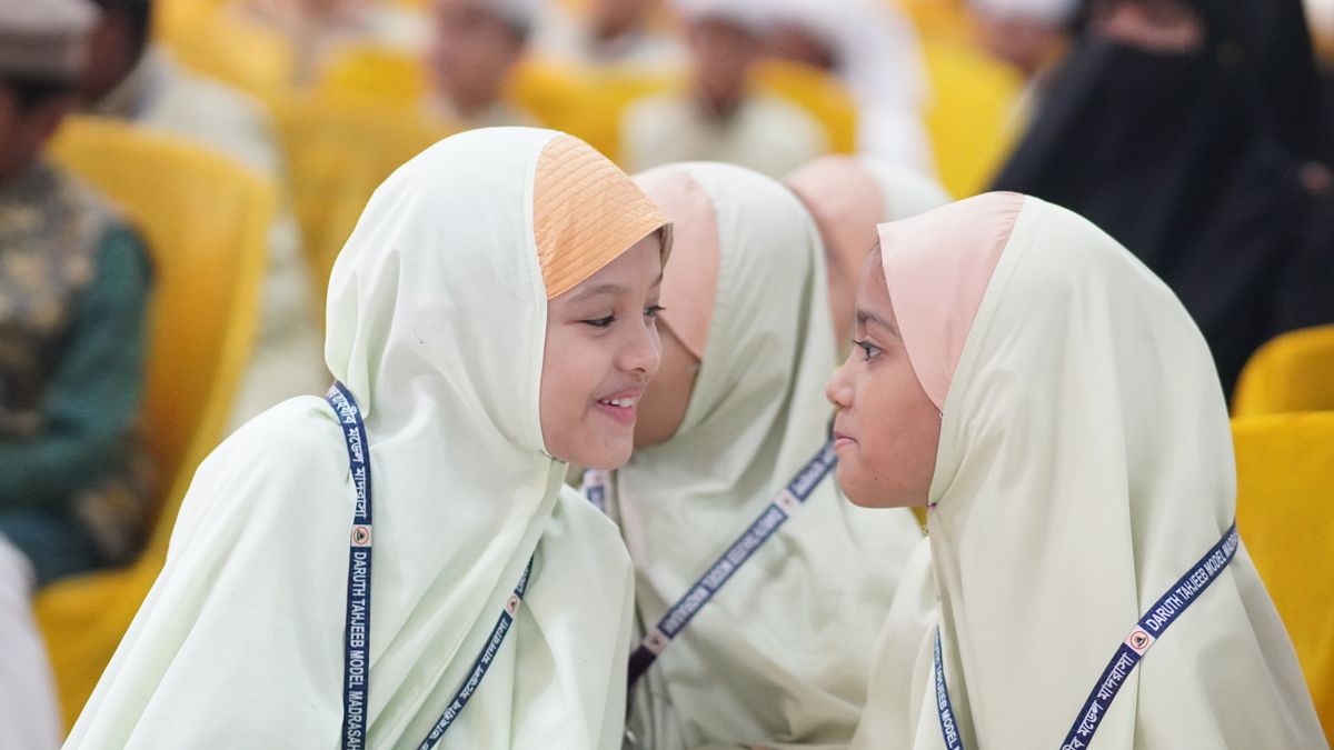 Two young girls engaging in conversation at a school event, wearing hijabs and ID lanyards.
