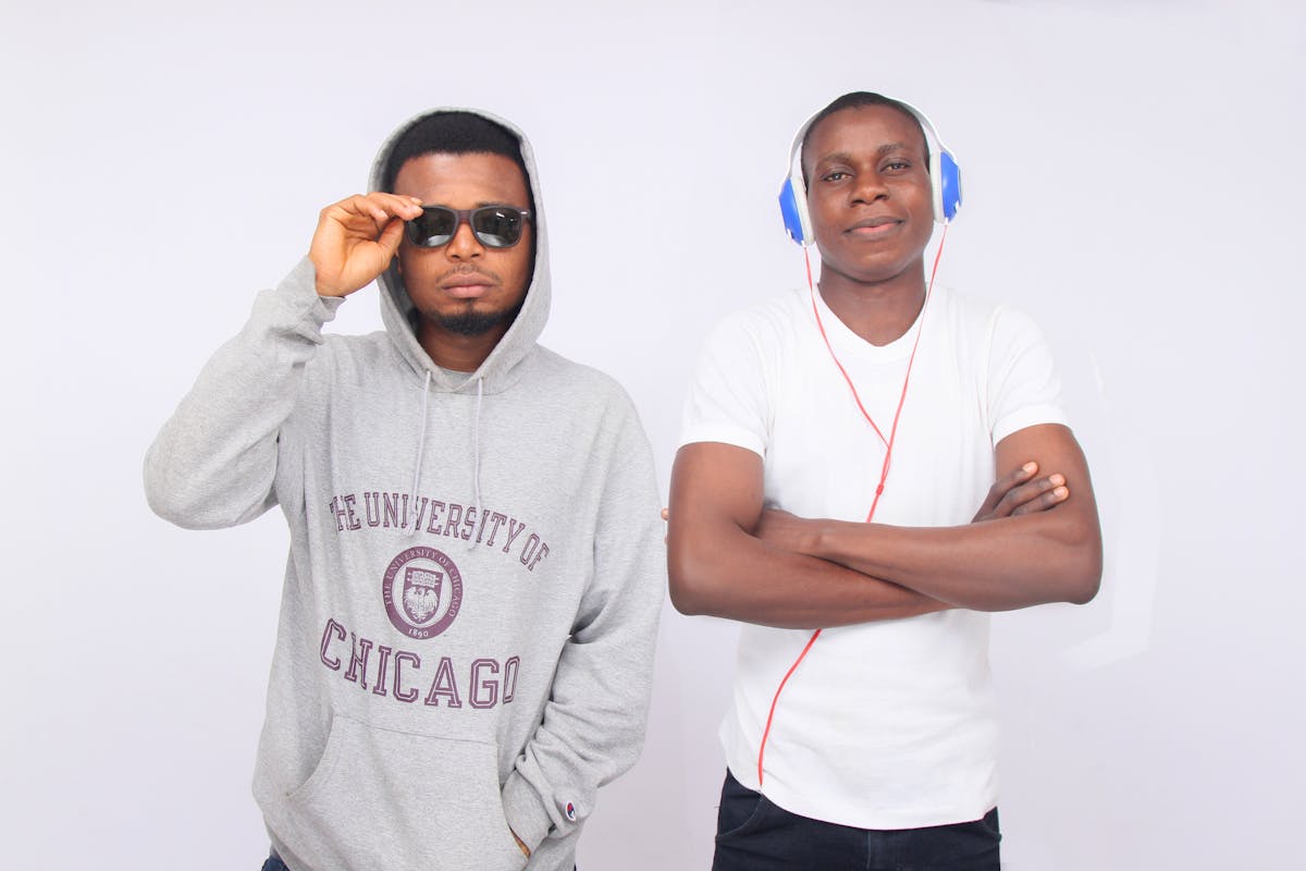 8 Men’s Fashion Choices That Scream “Notice Me Now.” 3 Two young men in casual attire, one wearing sunglasses and a hoodie, the other with headphones, against a white background.
