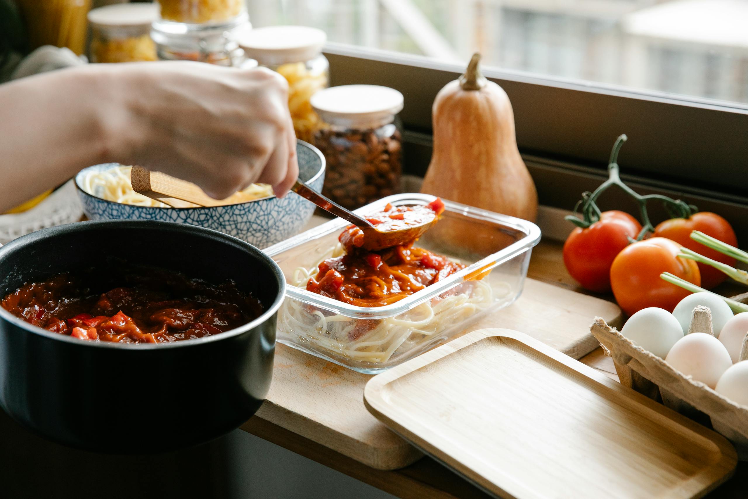 Unrecognizable cook pouring tasty sauce from saucepan into container with pasta while cooking at counter with ingredients in light kitchen