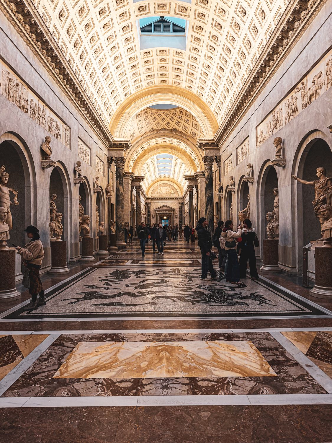 View of a grand museum hallway with classical sculptures and intricate ceilings, filled with visitors.