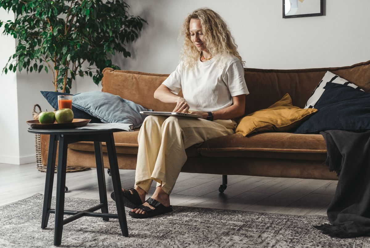 Woman sitting on couch using tablet, surrounded by cozy home decor and natural lighting.