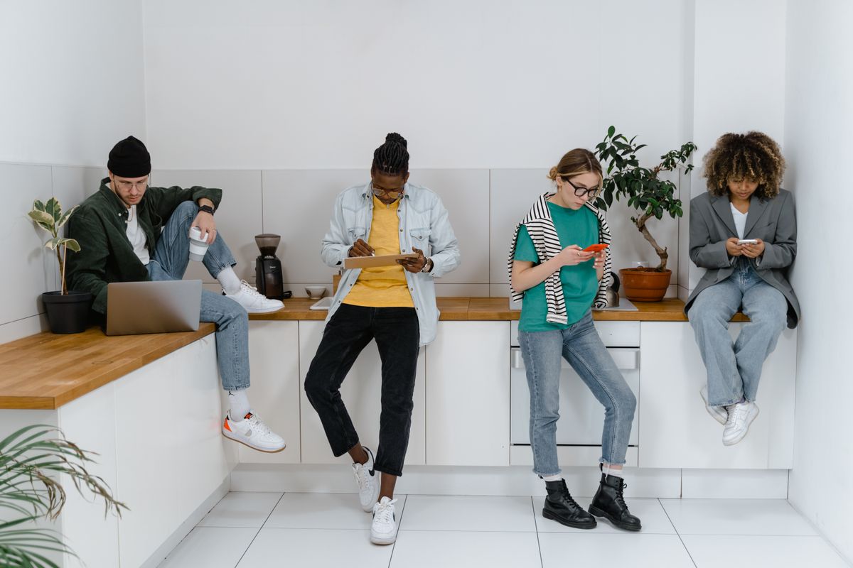 Gen Z Is The First Generation in 100 Years to Be Dumber Than Their Parents, Report Says 1 Young adults on digital devices in a modern office kitchen area, emphasizing diversity and teamwork.