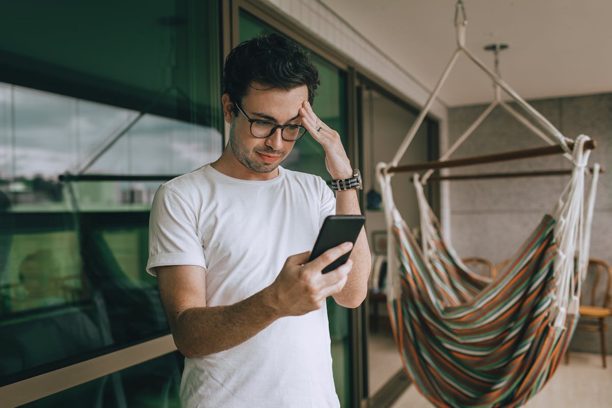 Young man with glasses checking his smartphone by a hammock indoors. Lifestyle and technology theme.