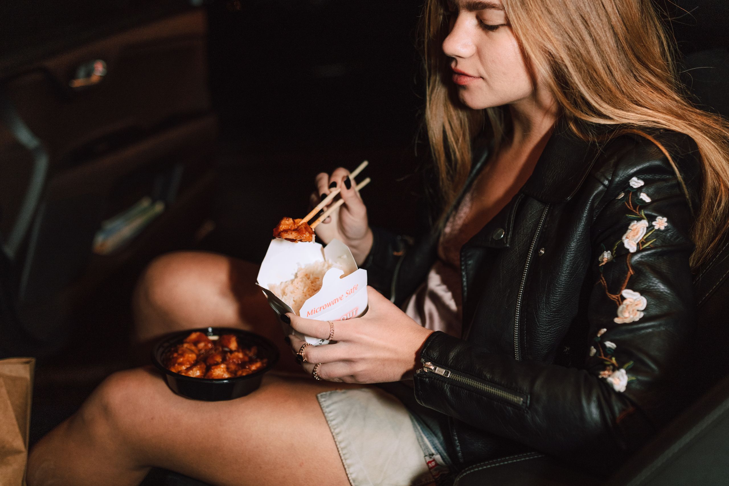 Young woman enjoying Asian takeout food while sitting in a car, wearing a black leather jacket.