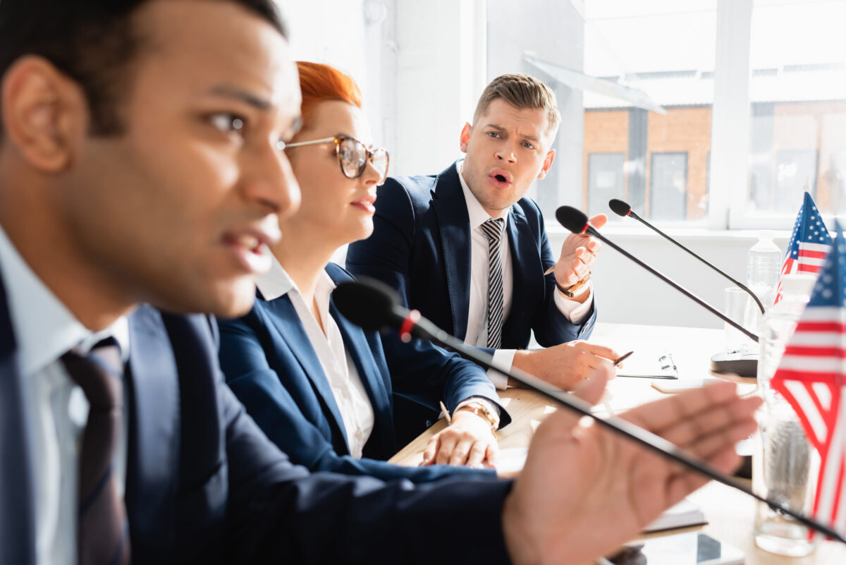 The No Getting Rich in Congress Act Takes Aim at Washington’s Wall Street Problem 2 Irritated politician with open mouth looking at colleague speaking in microphone, while sitting at table in boardroom on blurred foreground