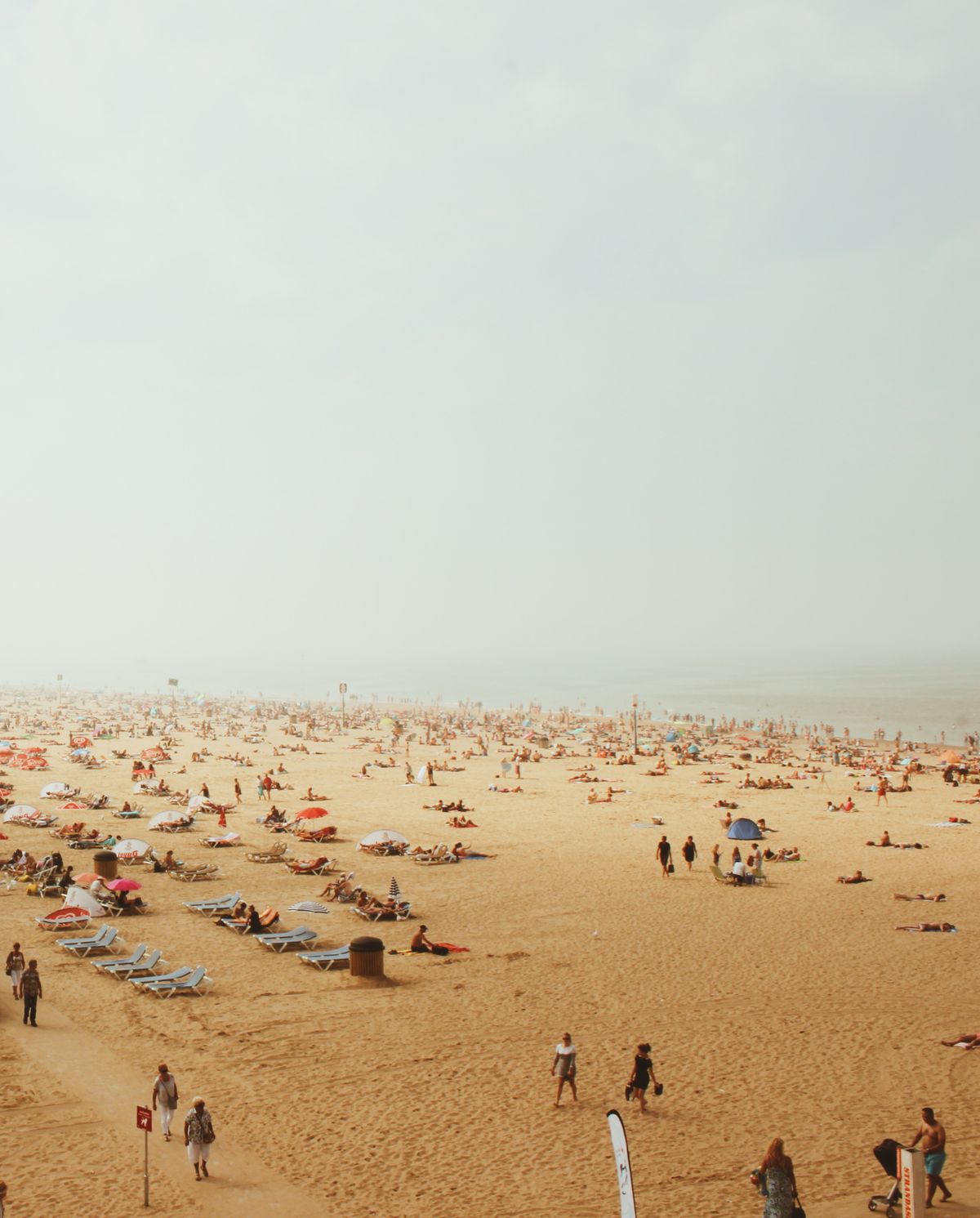 A bustling beach with many people sunbathing and relaxing on deckchairs under a hazy sky.