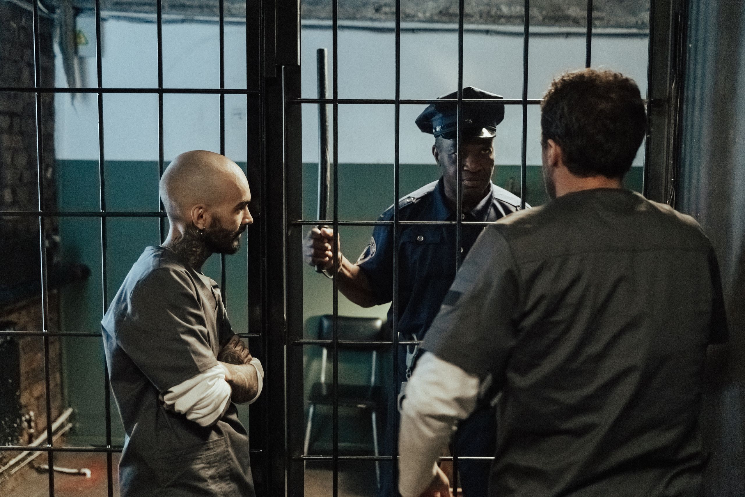 A prison guard interacts with two inmates inside a dimly lit jail cell.