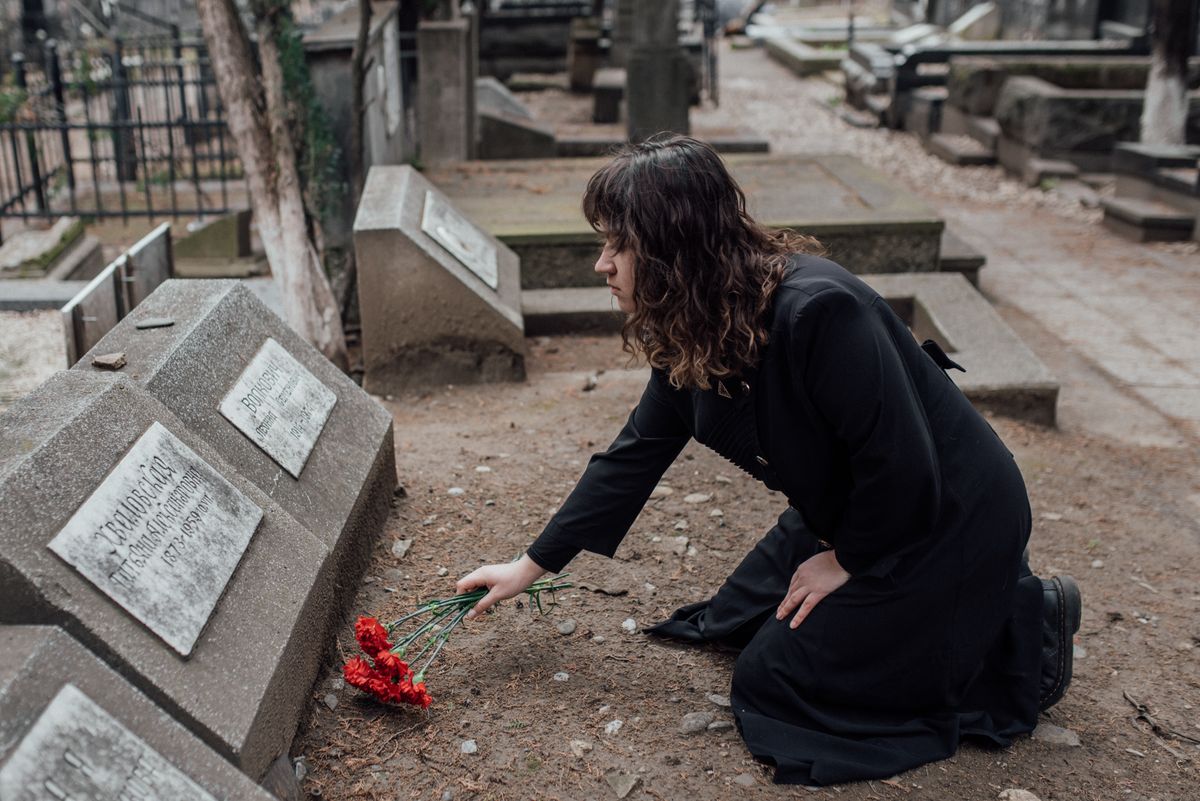 A woman in a black dress kneels by a tombstone with red flowers, expressing loss and remembrance.