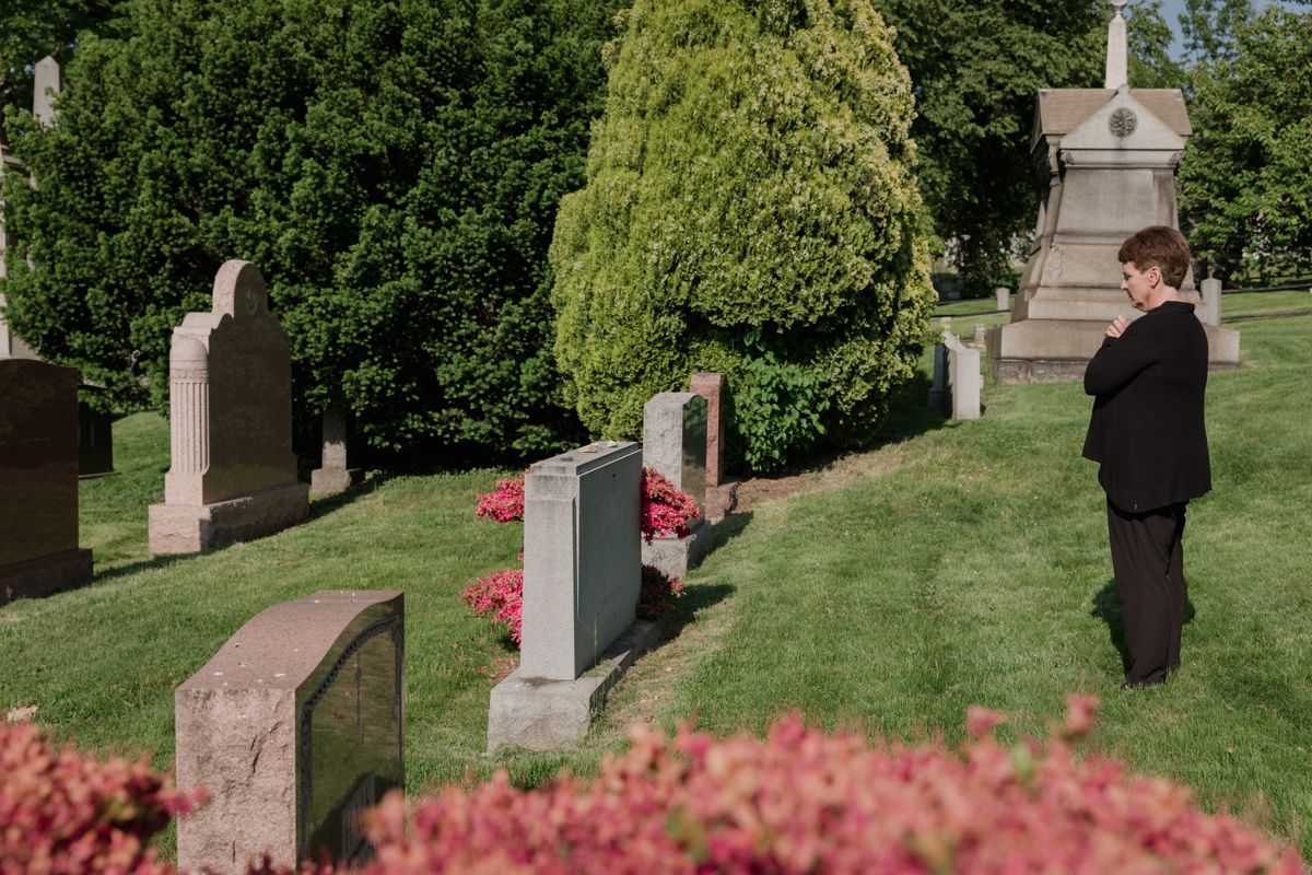A woman stands solemnly by gravestones in a tranquil cemetery, surrounded by greenery.