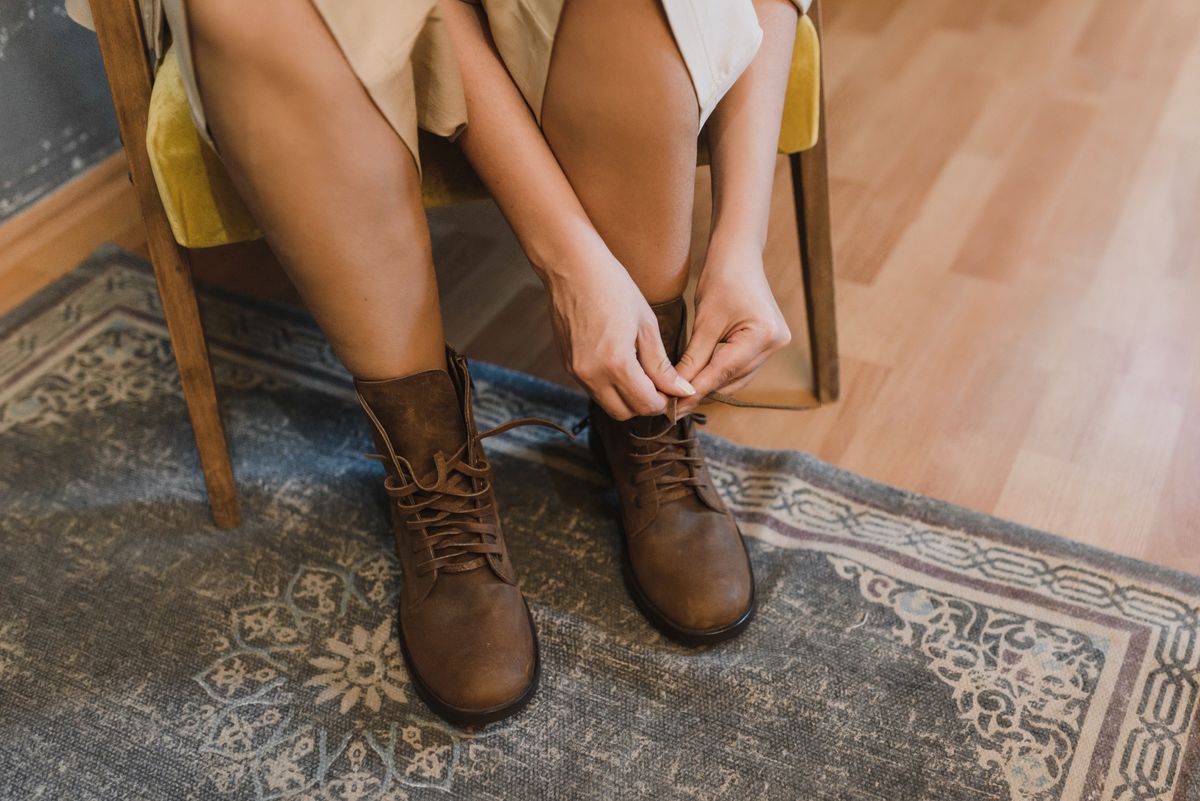 A woman tying brown leather boots while sitting on a chair, on a patterned rug.