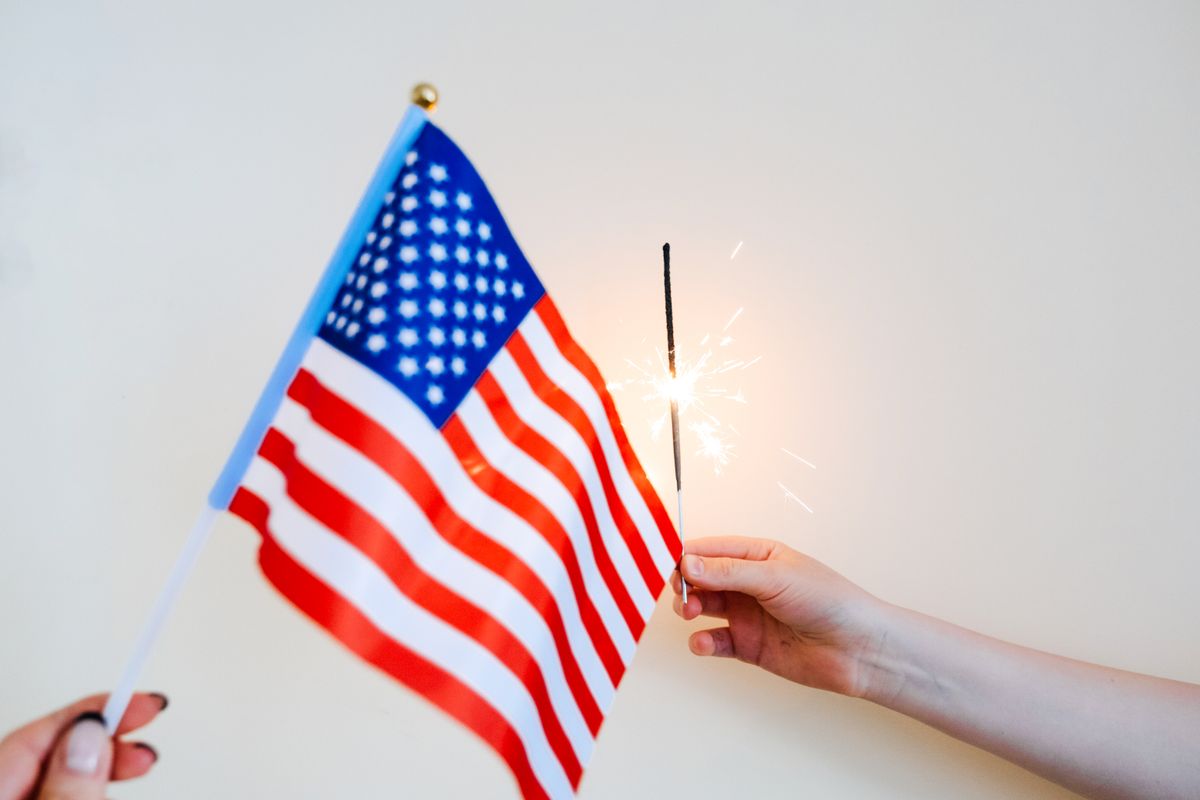 The Missing Trump Merchandise at the Great American State Fair and What It Really Signals 3 Close-up of hands holding an American flag and a sparkling sparkler on a neutral background.