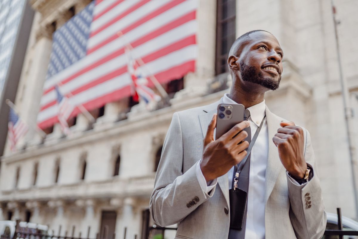 Confident businessman with phone in front of Wall Street building with American flags.