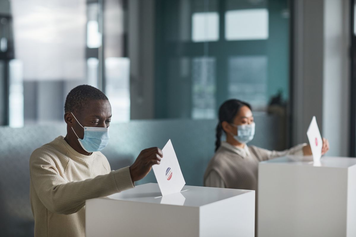 People wearing face masks cast ballots in a public indoor polling station with social distancing.
