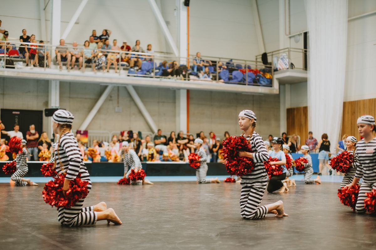 Performers in prisoner costumes and red pom-poms kneeling at a cheer competition indoors.