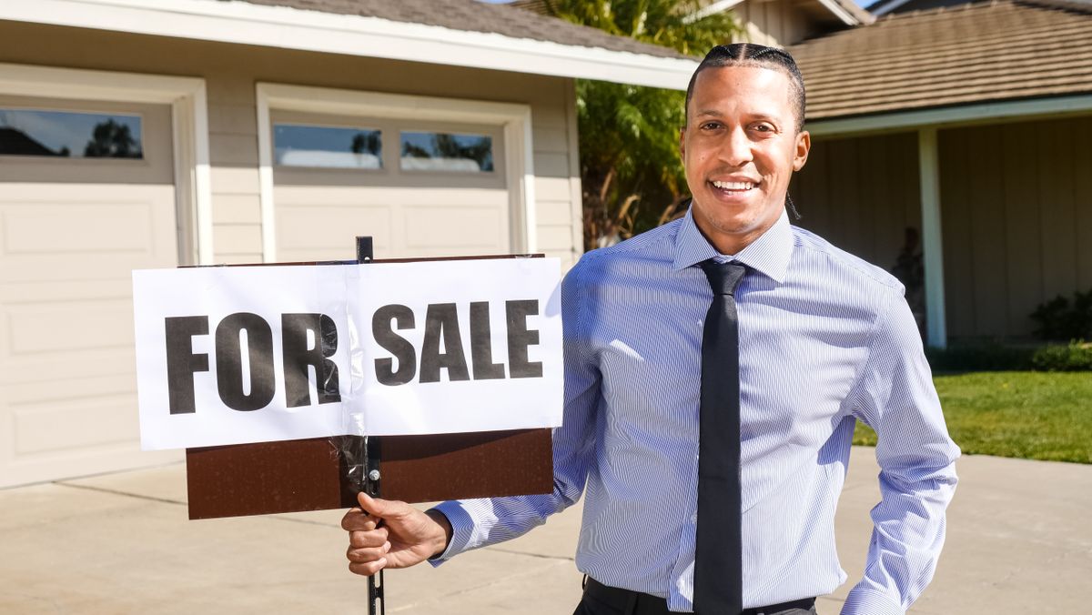 Smiling real estate agent with a for sale sign in front of a house.
