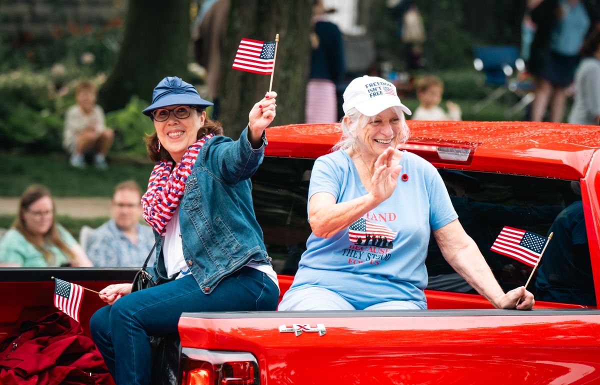 Two women enjoy a parade, proudly waving American flags from a red truck.